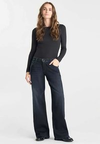 Haut noir à manches longues associé à un jean évasé foncé. Le denim présente un léger effet délavé, et la tenue comprend une poche avant ainsi que des passants pour ceinture.