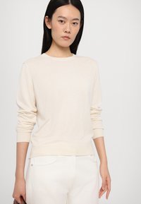 Maglione a maglia color crema con maniche lunghe e colletto rotondo. Texture morbida e polsini aderenti. Indossato con jeans bianchi. Design minimalista.