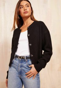 Black knitted cardigan with a round neck and five buttons. Paired with a white ribbed tank top and light blue high-waisted jeans.