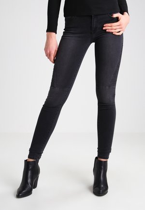 Femme portant un jean skinny côtelé noir et des bottines noires, posant avec une main sur la hanche devant un fond uni.