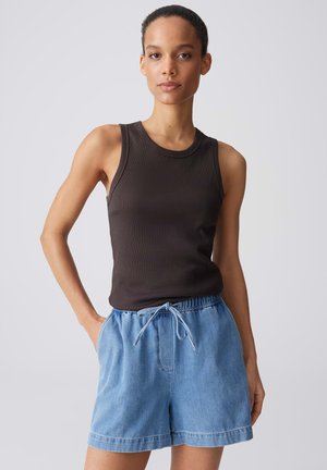 Vrouw met kort krullend haar die een mouwloos zwarte top en lichtblauwe spijkershorts met een koord in de taille draagt, staand met één hand in de zak.