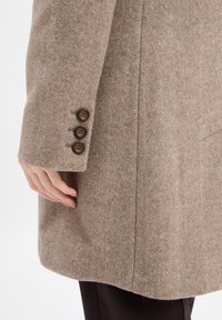 Una mano appoggiata accanto a una manica di un cappotto di lana beige a spina di pesce con tre bottoni marroni, abbinato a pantaloni scuri su uno sfondo bianco.