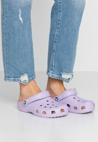 Cizme de culoare violet deschis, cu guri de ventilație, vârf rotund și curea la spate. Purtați cu blugi albastri destrămați, evidențiind încălțămintea casual.