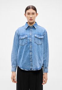 Svetlomodrá rifľová košeľa s golierom, dvoma náprsnými vreckami, zapínaním na gombíky a dlhými rukávmi. Má voľný strih a mierne zaoblený lem.