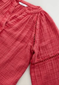 Blouse rouge texturée avec un décolleté en V, des boutons sur le devant et des détails en crochet sur les manches et l'ourlet. Fabriquée en tissu léger.