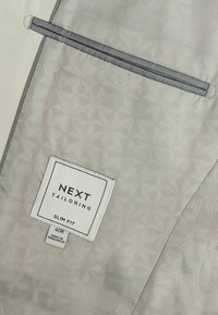 Intérieur en tissu gris avec un motif géométrique ; présente une étiquette slim fit et une petite poche entourée de gris, indiquant la marque de couture "NEXT".