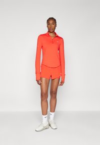 Korte mouwen, ritssluiting sporttop en shorts in feloranje. Gladde, elastische stof met witte Nike-logo-accenten. Sport sneakers met witte sokken.