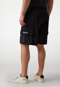 Schwarze Cargo-Shorts mit einem elastischen Bund, ausgestattet mit einer großen Seitentasche und GUESS-Branding. Getragen mit niedrigprofiligen Sneakers in hellen Farben.