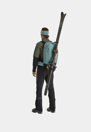 Back view of a person wearing a green backpack with ski straps, black pants, and a gray jacket, holding skis with black and orange details.
