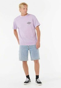 Camiseta de manga corta lila con un pequeño gráfico negro en el pecho, combinada con pantalones cortos de mezclilla azul claros y zapatillas negras con detalles blancos.