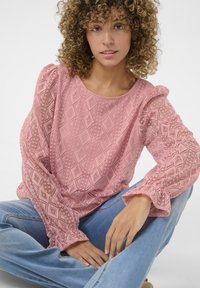 Pink lace blouse with geometric patterns, puffed sleeves, and ruffled cuffs; worn with light blue jeans. Soft, textured fabric.