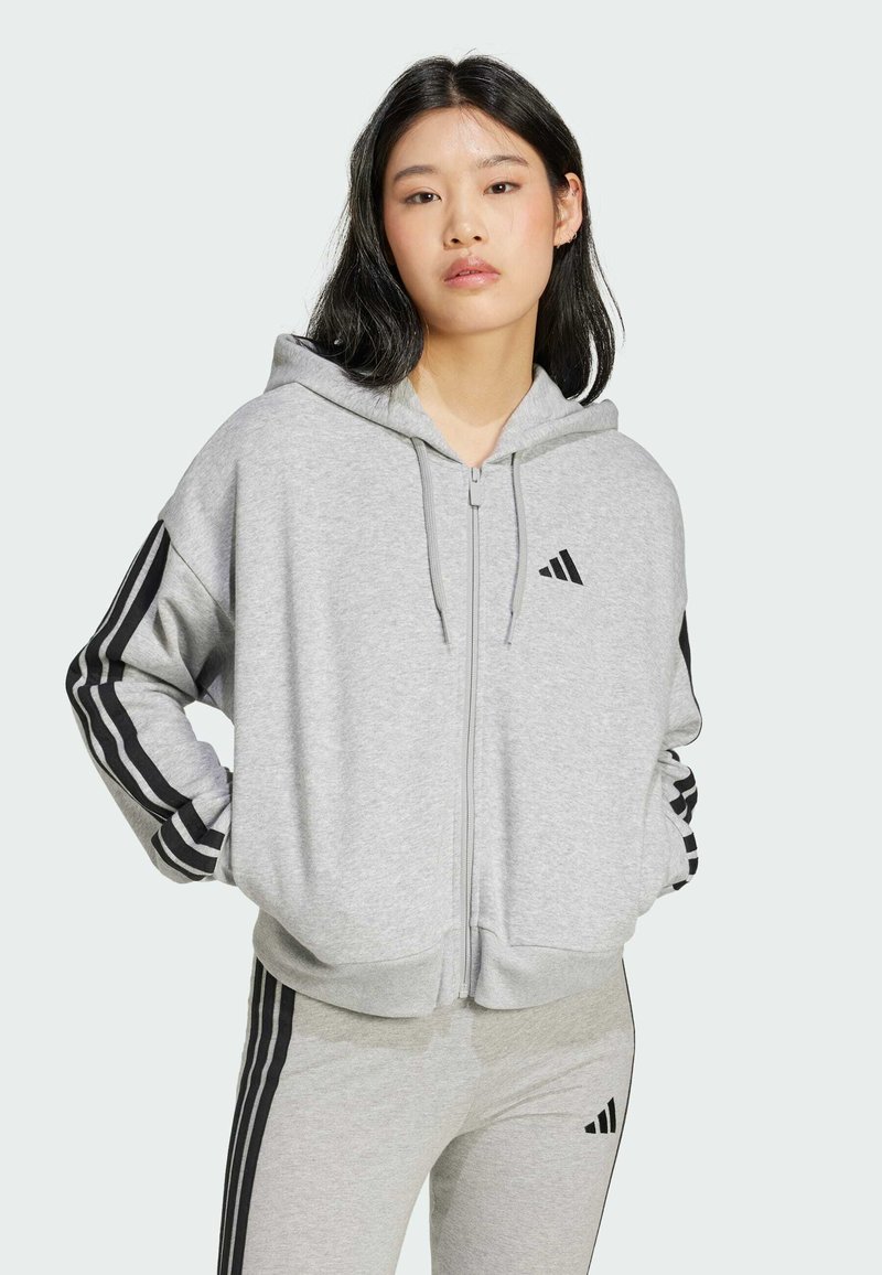 Sweatshirt cinza com fecho, feito de um tecido macio, apresentando riscas laterais pretas e um pequeno logo no peito. Inclui um capuz espaçoso e bolsos frontais.
