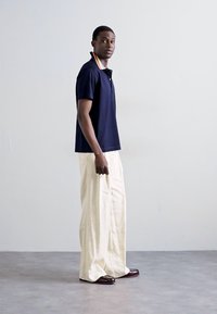 Polo shirt marine avec un col rayé, associé à un pantalon large crème et des chaussures marron foncé, se tenant sur un sol gris contre un fond uni.