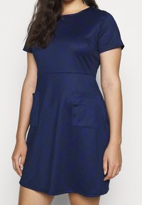 Abito blu navy in un tessuto morbido con maniche corte, caratterizzato da un motivo a rombi viola e due tasche frontali.