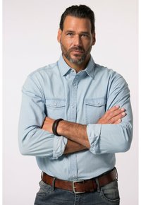 Camicia in denim azzurro chiaro con tasche sul petto abbottinate, maniche lunghe arrotolate ai polsini e una cintura in pelle marrone. Uno sfondo neutro.