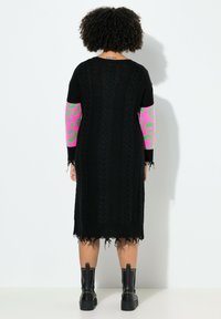 Schwarzes Strickkleid mit langen Ärmeln im pinken und grünen Tierprint. Die Kanten sind fransig verarbeitet; getragen mit schwarzen, mit Nieten besetzten Stiefeletten.