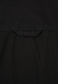 Tessuto nero con una texture liscia, caratterizzato da un'etichetta cucita al centro che mostra il nome del marchio. Design semplice e minimalista.