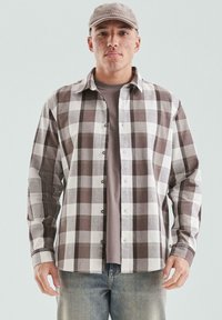 Młody mężczyzna z piegami, ubrany w brązową i białą koszulę w kratę, taupe t-shirt, beżową czapkę oraz wyblakłe niebieskie dżinsy, stoi na tle jednolitym.