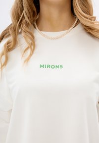 Biała bluza z bawełny, z zielonym haftem "MIRONS" na klatce piersiowej. Naszyjnik z małych pereł widoczny wokół szyi.