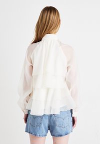 Hvit lagdelt bluse med gjennomsiktig stoff, volanger og lange ermer. Kombinert med lyse denimshorts med knappelukking.