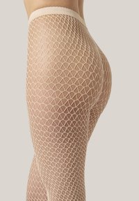 Närbild av en person som bär köttfärgade nätstrumpbyxor med ett diamantmönster mot en enkel ljus bakgrund.