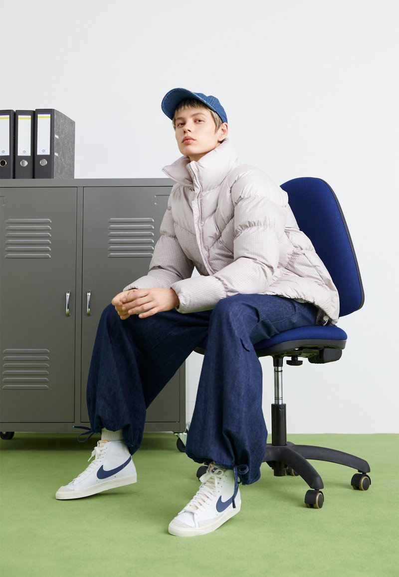 Doudoune beige clair, pantalon large en denim bleu, baskets montantes blanches avec des accents marins, assis sur une chaise de bureau bleue.