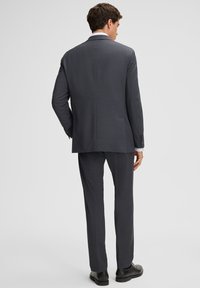 Mężczyzna stojący tyłem w ciemnoszarym garniturze, białej koszuli i czarnych butach wizytowych na jednolitym jasnym tle.