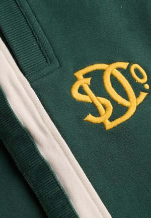 Zielona tkanina z żółto haftowanymi splecionymi literami "S" i "C" obok białego paska i prążkowanego detalu krawędzi.