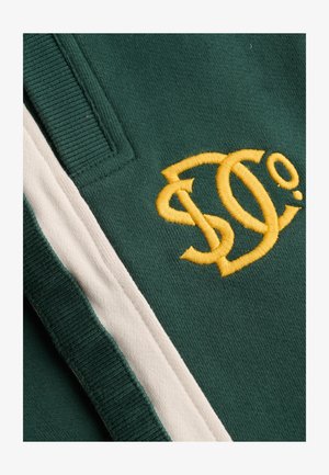 Zielona tkanina z żółto haftowanymi splecionymi literami "S" i "C" obok białego paska i prążkowanego detalu krawędzi.