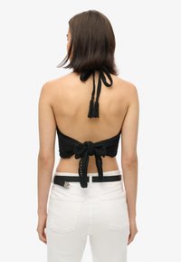 Top de crochet negro sin mangas con cierre de lazo y acentos de borlas. Lleva puesto sobre unos jeans blancos de cintura alta, mostrando un diseño de espalda descubierta.