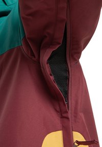 Chaqueta color marrón y verde azulado con un bolsillo interior de malla en el lateral, que presenta acentos amarillos y detalles de costura en contraste.