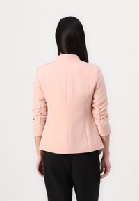 Roze op maat gemaakte blazer met een hoge kraag, gerimpelde mouwen en een gestructureerde pasvorm, gemaakt van gladde stof, gecombineerd met zwarte broek.