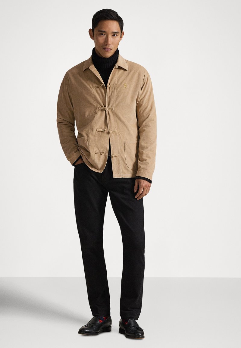 Mann trägt eine beige Jacke über einem schwarzen Rollkragenpullover, schwarze Hose und schwarze Loafers, steht vor einem schlichten weißen Hintergrund.