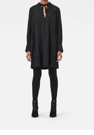 Kvinde iført en løs sort kjole med lange ærmer og bindebånd ved halsen, sorte leggings og sorte ankelstøvler med lynlås foran.