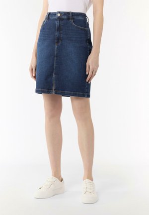 Kvinde iført en knælængde blå denimnederdel med forreste sømme, hvide sneakers og en hvid top, der er stukket ind, stående mod en ensfarvet hvid baggrund.