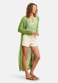 Langer, offener Strickcardigan in Grün über einem weißen, gemusterten Hemd, kombiniert mit beigen Shorts und braunen Sandalen. Strukturstrick und leichtes Material.