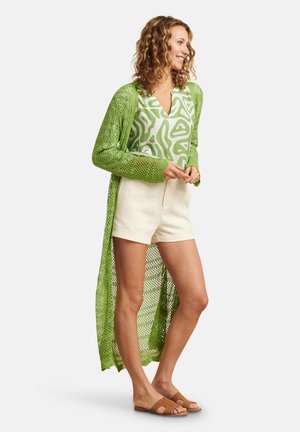 Lange, opengewerkte groene cardigan over een witte shirt met patroon, gecombineerd met beige shorts en bruine sandalen. Getextureerd breiwerk en lichtgewicht stof.