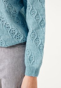 Lichtblauwe gebreide trui met een ruitpatroon en opengewerkte details, ribbelmanchetten en een losse pasvorm. De textuur is zacht en luchtig.