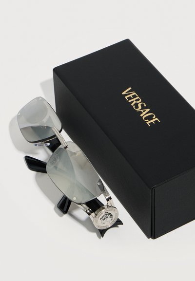 Μαύρα γυαλιά ηλίου Versace με ασημένιο πλαίσιο και έμβλημα Medusa που ακουμπούν δίπλα σε μια μαύρη θήκη Versace με χρυσά γράμματα πάνω σε λευκή επιφάνεια.