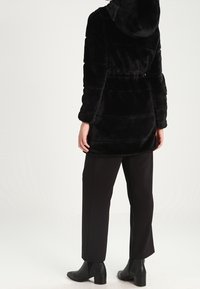 Černý kabát z umělé kožešiny s kapucí, s vypasovaným pasem, horizontálním prošíváním a dlouhými rukávy, doplněný o rovné černé kalhoty a kotníkové boty.