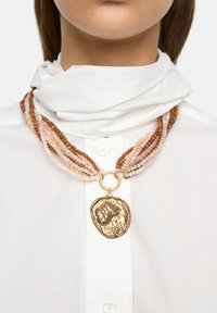 Naszyjnik z wieloma rzędami beżowych i brązowych koralików z teksturowanym złotym wisiorkiem zawieszonym na metalowym kółku. Na tle białej koszuli.