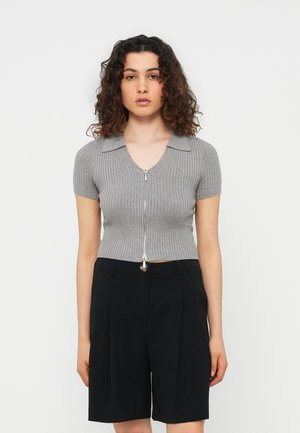 Femme aux cheveux bouclés portant un haut gris côtelé moulant à manches courtes avec fermeture éclair à l'avant et un short noir taille haute arrivant aux genoux.