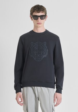 Jeune homme portant un sweat-shirt noir avec un visage de tigre en relief, un pantalon cargo gris clair et de grandes lunettes de soleil noires, se tenant devant un fond uni.