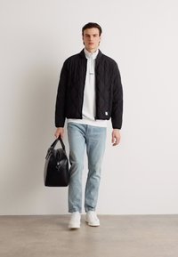 Chaqueta bomber negra acolchada con bolsillos con cremallera, camisa blanca sin cuello debajo, jeans de color azul claro y bolso negro. Zapatillas blancas completan el look.