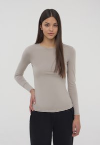 Maglietta a maniche lunghe di un leggero beige, realizzata in tessuto morbido, con scollatura rotonda e design aderente, indossata con pantaloni neri, con cuciture e dettagli minimi.