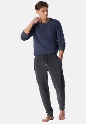 Jonge man met een blauw gestreept shirt met lange mouwen en donkergrijze joggingbroek, staat op blote voeten met één hand in de zak en de andere die zijn nek raakt.