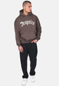 Brun hætte sweatshirt med "Dropsize" i hvid tekst, frontlomme, ribbede manchetter, kombineret med sorte bukser og hvide sneakers.
