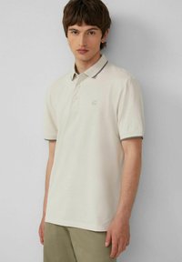 Beige Polohemd aus Baumwolle mit einer Knopfleiste mit drei Knöpfen, kurzen Ärmeln und kontrastierendem Kragen sowie Ärmelsaum. Dezentes Logodesign.