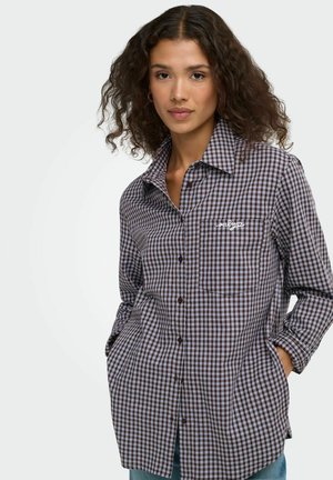 Femme aux cheveux bouclés portant une chemise à carreaux marron et bleu boutonnée, les mains dans les poches, face à un fond clair.