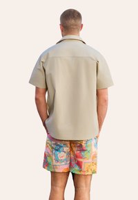 Korte mouw, beige knoopshirt met een ontspannen pasvorm, gedragen met kleurrijke, patroon zwembroek met bloemmotieven en levendige kleuren.